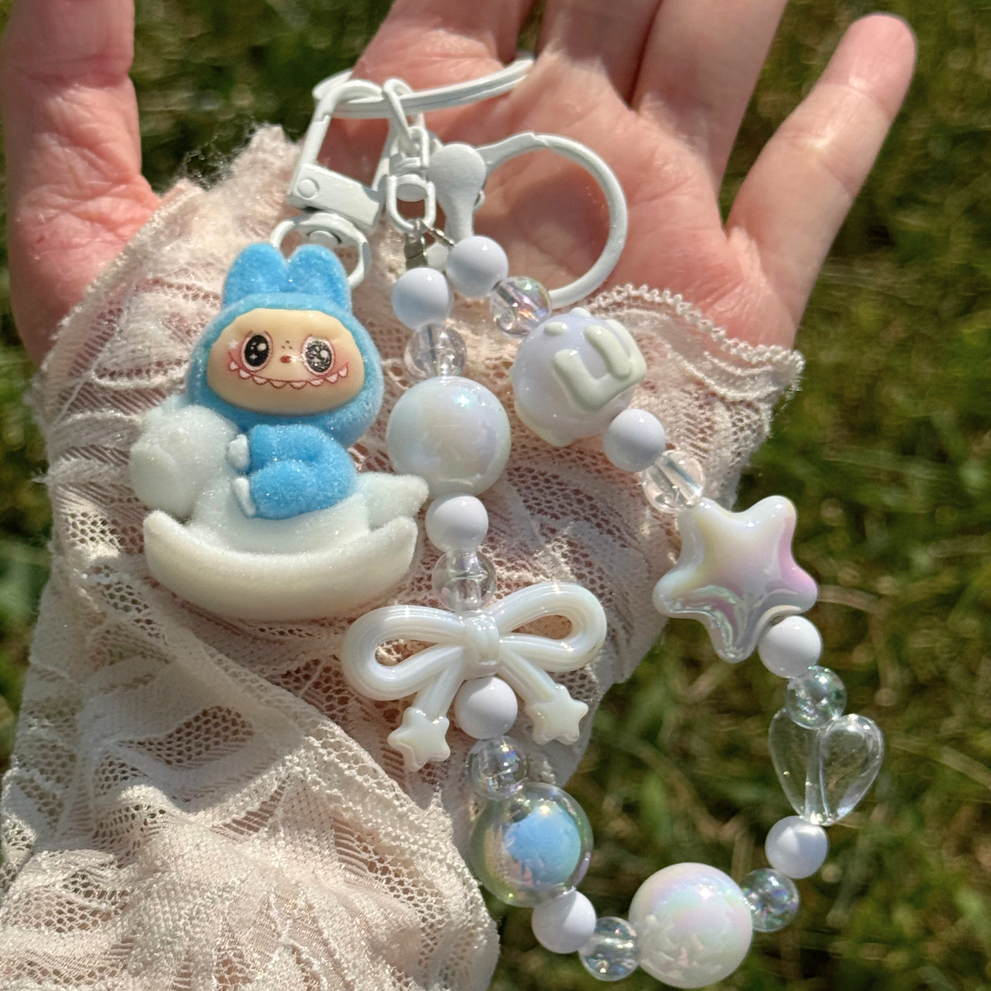 A pastel beaded keychain bracelet with a blue bunny plush rocking chair charm, white beads, iridescent star and pearl charms, and a white bow, displayed on a white tray outdoors with grass visible in the background.