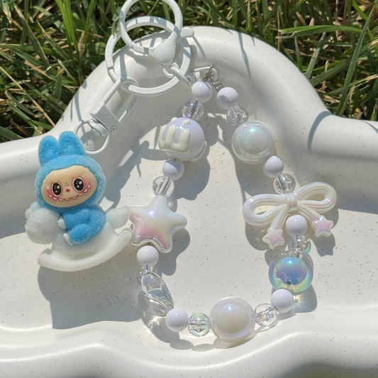 A pastel beaded keychain bracelet with a blue bunny plush rocking chair charm, white beads, iridescent star and pearl charms, and a white bow, displayed on a white tray outdoors with grass visible in the background.