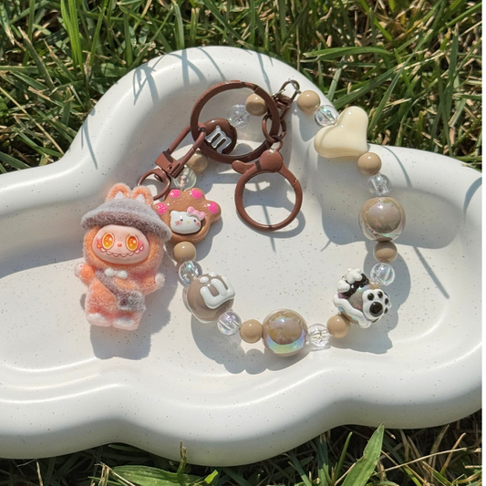 A beaded charm bracelet with a cute orange plush character, a small pink bear charm, a brown metal key ring, and decorative beads, displayed on a white cloud-shaped tray set outdoors on green grass.